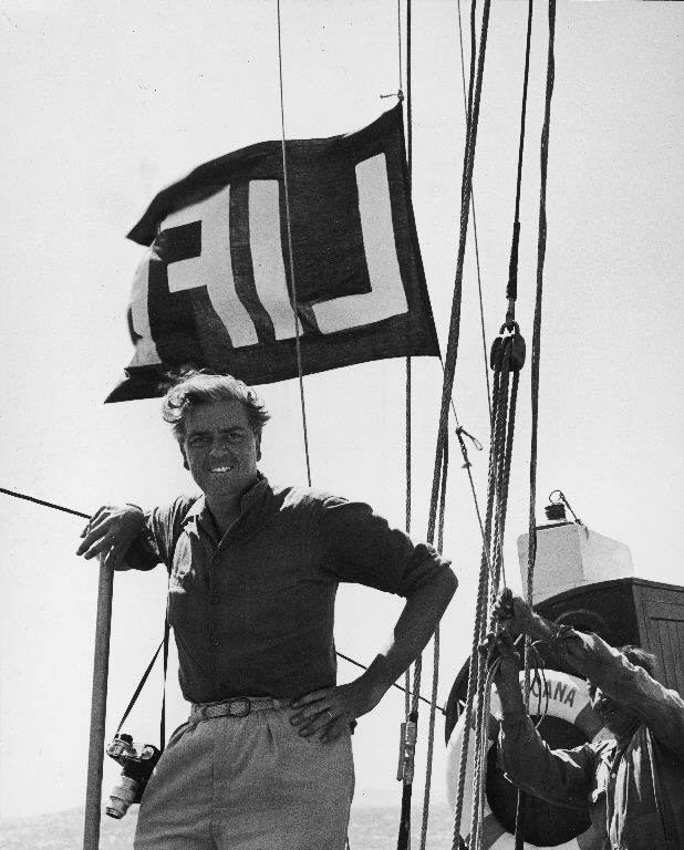 Photographer John Phillips aboard boat posing in front of LIFE flag he hoisted in hopes of getting pictures of a Greek royal cruise from which press was barred. (Photo by John Phillips/The LIFE Picture Collection © Meredith Corporation)
