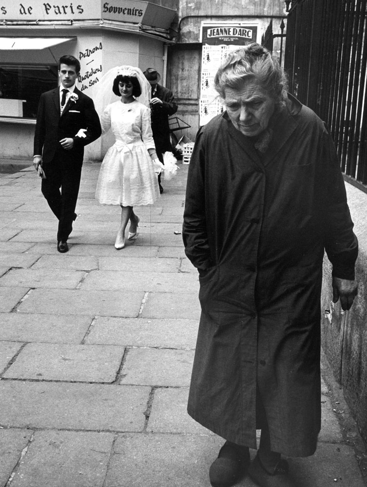 Elderly woman walking along street while bride and groom walk behind, Paris, 1963.